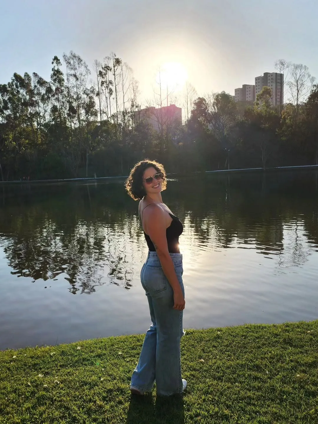Jacqueline Junqueira sorrindo de óculos escuros à beira de um lago, num fim de tarde de sol