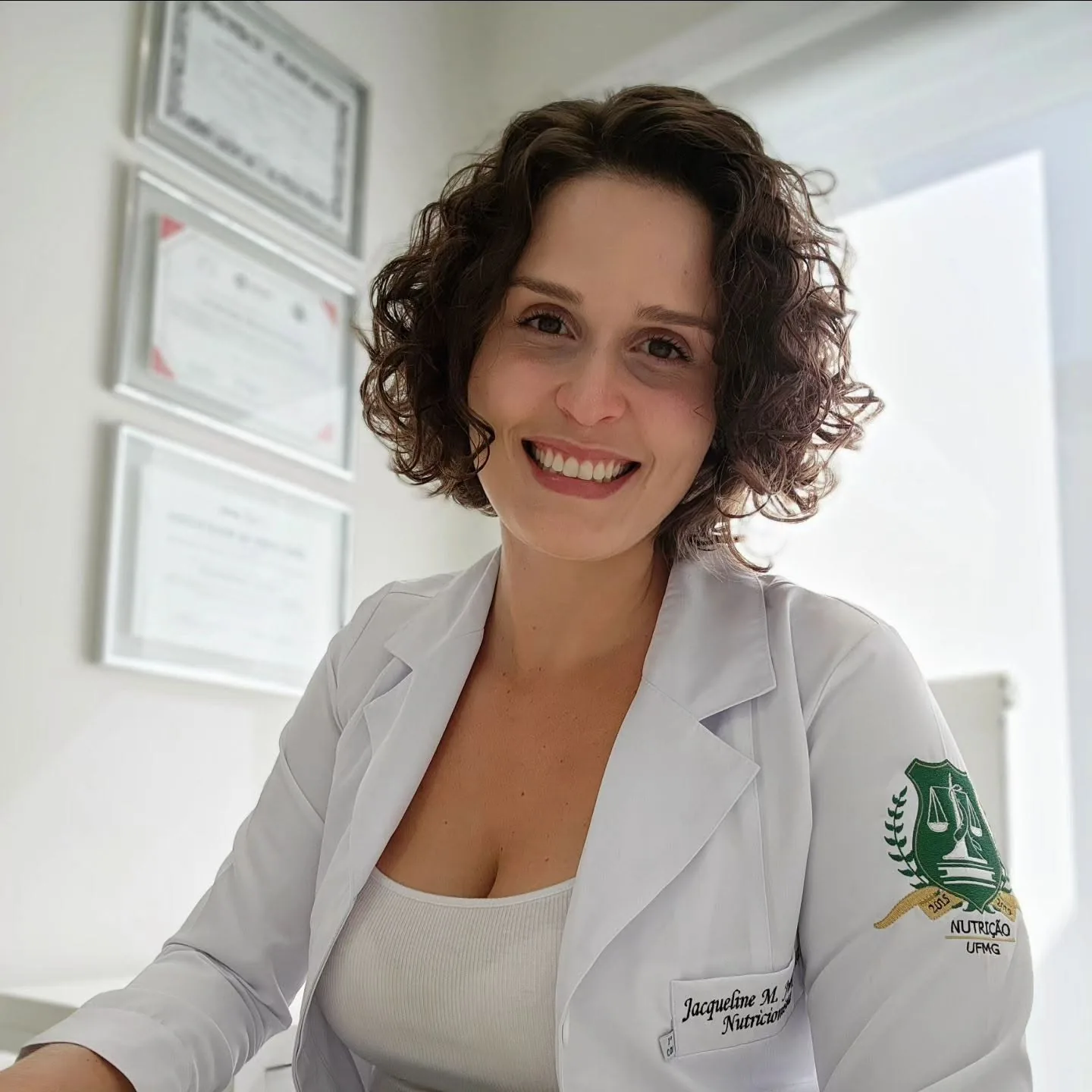 Jacqueline Junqueira sorrindo, de jaleco branco com o brasão da Nutrição UFMG, no consultório com certificados na parede